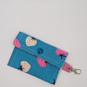 Könnte beinhalten: Blaues Stoffportemonnaie mit einem Muster aus rosa und cremefarbenen Erdbeeren. Es hat einen Klappenverschluss mit Knopf und einen passenden Riemen mit silberfarbenem Verschluss. Das Innere ist weiß.