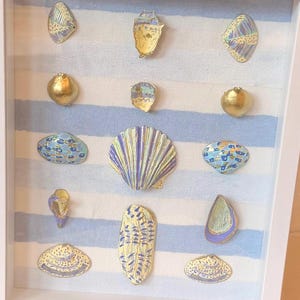 May include: A white shadow box frame containing a collection of twelve decorative seashells arranged in a grid. The background is a light blue and white striped fabric. The shells feature various painted patterns in gold, blue, and purple tones.