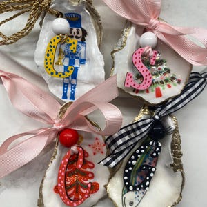 Puede incluir: Cuatro adornos de concha de ostra con diseños pintados y cintas rosas. Uno presenta un cascanueces, otro una letra "S" rosa y un árbol de Navidad, un tercero un árbol de Navidad rojo y el último un pez.