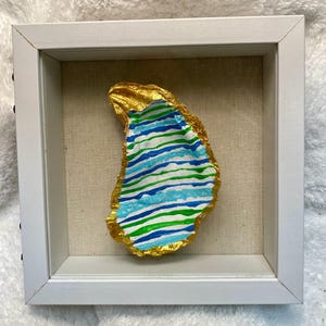 May include: A framed, painted oyster shell. The shell is painted with horizontal stripes of blue and green, with a gold-colored edge. The shell is set against a neutral background within a white frame.