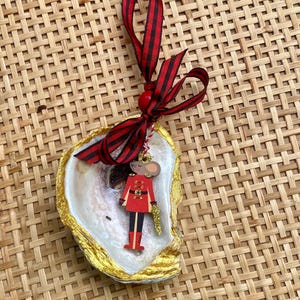 Peut inclure: Ornement de Noël avec une souris en bois en uniforme rouge, tenant un bâton doré, placé dans une coquille d'huître dorée. L'ornement est suspendu à un ruban rayé rouge et noir.