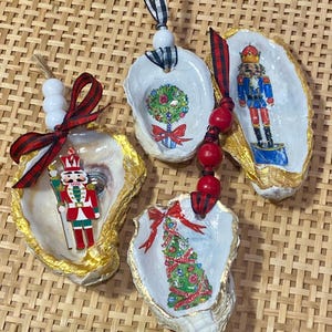 May include: Four Christmas ornaments made from oyster shells. Each shell is painted with a different holiday design, including a nutcracker, a Christmas tree, and a wreath. The shells are trimmed with gold paint and have ribbons for hanging.