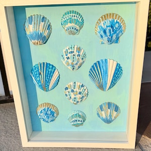 May include: A framed artwork featuring nine painted seashells in shades of blue, white, and gold. The shells are arranged on a light blue and green background within a white frame. The artwork is a decorative piece.