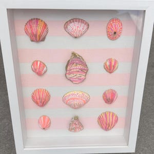 Könnte beinhalten: Ein weißer Schattenfugenrahmen mit zwölf Muscheln, die in einem Raster auf einem hellrosa und weiß gestreiften Stoffhintergrund angeordnet sind. Jede Muschel ist individuell mit leuchtend rosa, orangefarbenen und goldenen Streifen bemalt.