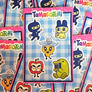 May include: A collection of Tamagotchi sticker sheets featuring various cartoon characters. The stickers include a ninja, a bunny with a drink, and other colorful characters. The sheets have a pink border and are set against a blue and white checkered background.