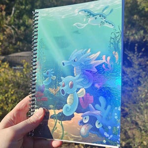 May include: Spiral-bound notebook featuring an underwater scene with various cartoon seahorse and sea creature illustrations in shades of blue, green, and purple. The cover has a holographic, iridescent finish, and the notebook is held in a person's hand.
