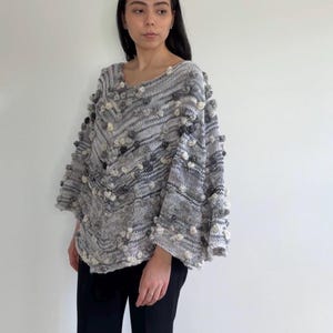 May include: A grey and white knit poncho featuring a chevron pattern and scattered pom-pom embellishments in varying shades of grey and cream. The garment has a wide, loose-fitting silhouette with a V-neckline and textured knit construction.