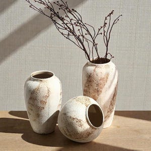 Puede incluir: Tres jarrones de cerámica blanquecina con acabado texturizado y detalles marrones. Un jarrón es esférico y los otros dos son altos y cilíndricos. El jarrón más alto contiene ramas marrones. Los jarrones están colocados sobre una superficie de madera.