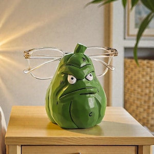 Puede incluir: Un soporte para gafas verde y caricaturesco con forma de verdura gruñona con ojos blancos y una expresión severa. Las gafas con montura transparente descansan sobre el soporte. El soporte está sobre una mesita de noche de madera.