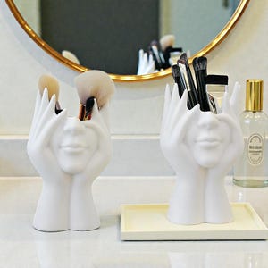 Puede incluir: Dos soportes blancos de cerámica para brochas de maquillaje, cada uno con forma de rostro con las manos cubriendo los ojos. Uno contiene brochas beige y marrones, el otro brochas negras. Un espejo con marco dorado está en el fondo.