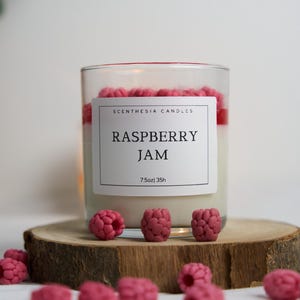 Może przedstawiać: Przezroczysta szklana świeca z białym wnętrzem i czerwoną warstwą wierzchnią, oznaczona jako "Raspberry Jam" przez "Scenthesia Candles". Świeca stoi na drewnianym plastrze, otoczona dekoracyjnymi woskowymi kawałkami w kształcie malin. Świeca ma 213g i czas palenia 35h.