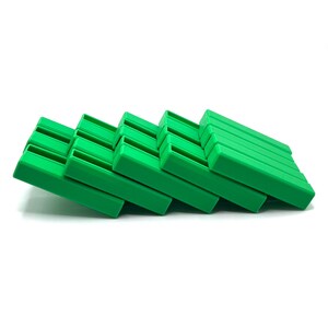 May include: A stack of bright green plastic soap dishes, each with a rectangular shape and a series of parallel ridges on the top surface. The dishes are arranged in a diagonal line against a white background.