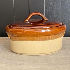 May include: A two-tone ceramic casserole dish with a lid. The lid and rim are a glossy brown, while the base is a light tan. The dish has a handle on the lid and is suitable for baking and serving.