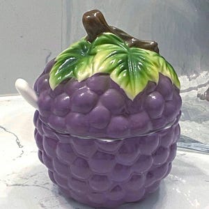 May include: A ceramic jar in the shape of a bunch of purple grapes, with a green and yellow leaf design on the lid. The lid has a brown stem. A small white spoon is on the side of the jar.