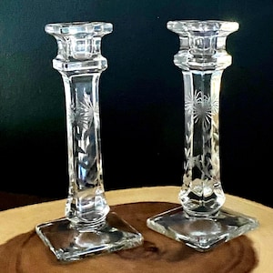 May include: A pair of clear glass candle holders with etched floral patterns. Each has a square base, a slender stem, and a square top. The candle holders are presented on a wooden surface against a dark backdrop.