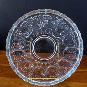 Puede incluir: Una fuente de vidrio transparente con un patrón floral decorativo. La fuente tiene un borde festoneado y está sostenida por un soporte de metal dorado. El centro de la fuente es un círculo sólido, y la fuente está sobre una superficie de madera.