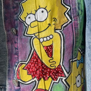 K&ouml;nnte beinhalten: Jeansjacke mit handgemaltem Cartoon-Charakter-Design in Gelb, Rot und Wei&szlig;. Die Jacke hat silberne Kn&ouml;pfe und einen Batik-Effekt im Hintergrund.