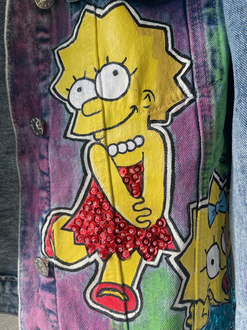 K&ouml;nnte beinhalten: Jeansjacke mit handgemaltem Cartoon-Charakter-Design in Gelb, Rot und Wei&szlig;. Die Jacke hat silberne Kn&ouml;pfe und einen Batik-Effekt im Hintergrund.