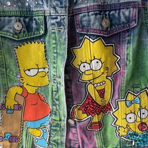 K&ouml;nnte beinhalten: Jeansjacke mit handgemalten Bildern von Bart, Lisa und Maggie Simpson. Die Jacke hat eine Knopfleiste, Taschen und ist mit Strasssteinen verziert. Der Hintergrund hat einen Batik-Effekt mit gr&uuml;nen, lila und blauen Farbt&ouml;nen.