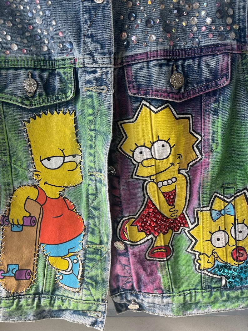 K&ouml;nnte beinhalten: Jeansjacke mit handgemalten Bildern von Bart, Lisa und Maggie Simpson. Die Jacke hat eine Knopfleiste, Taschen und ist mit Strasssteinen verziert. Der Hintergrund hat einen Batik-Effekt mit gr&uuml;nen, lila und blauen Farbt&ouml;nen.