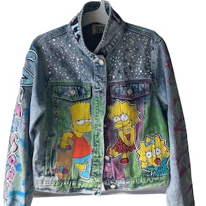 May include: A light-wash denim jacket adorned with colorful cartoon characters and sparkling embellishments. The jacket features a collage of painted designs, including Bart Simpson with a skateboard and Lisa Simpson with Maggie.