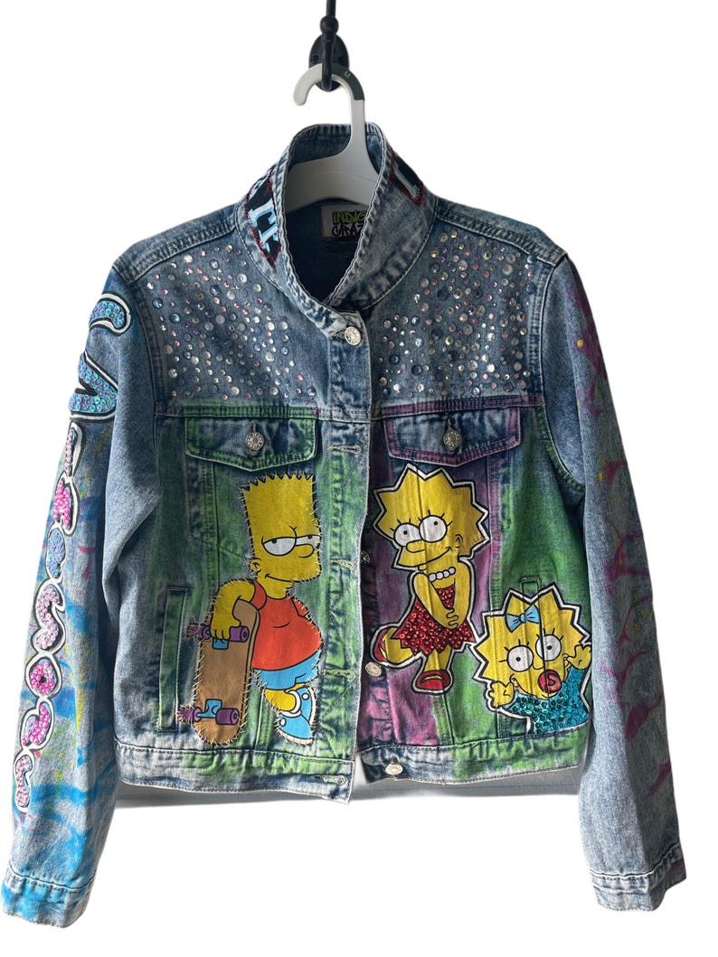 K&ouml;nnte beinhalten: Eine hellblaue Jeansjacke mit bunten Cartoonfiguren und glitzernden Verzierungen. Die Jacke hat bemalte Designs, darunter Bart Simpson mit einem Skateboard und Lisa Simpson mit Maggie.