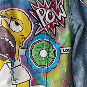 K&ouml;nnte beinhalten: Jeansjacke mit handgemalten Cartoon-Grafiken. Das Design umfasst eine gelbe Figur, einen gr&uuml;nen Donut und das Wort "POW" in einem roten Burst. Der Hintergrund hat einen Batik-Effekt mit dem Wort "LOVE".