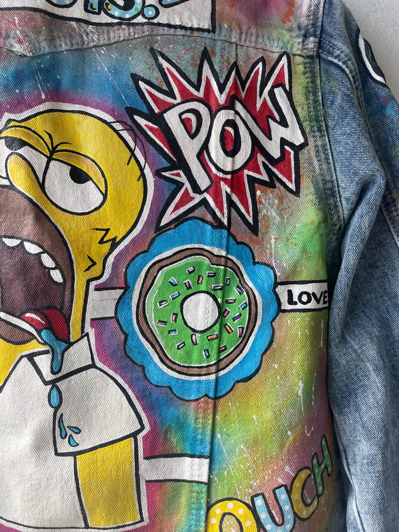 K&ouml;nnte beinhalten: Jeansjacke mit handgemalten Cartoon-Grafiken. Das Design umfasst eine gelbe Figur, einen gr&uuml;nen Donut und das Wort "POW" in einem roten Burst. Der Hintergrund hat einen Batik-Effekt mit dem Wort "LOVE".