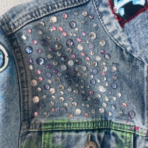 K&ouml;nnte beinhalten: Nahaufnahme einer hellblauen Jeansjacke, verziert mit verschieden gro&szlig;en, irisierenden Strasssteinen. Die Jacke hat einen Aufn&auml;her mit blauem Text und roten N&auml;hten. Die Tasche hat ein gr&uuml;n-gelbes Design und einen silbernen Knopf mit der Aufschrift "JEANS".