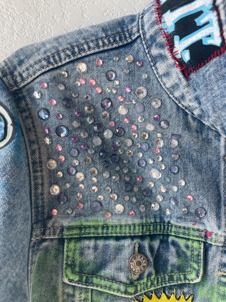 K&ouml;nnte beinhalten: Nahaufnahme einer hellblauen Jeansjacke, verziert mit verschieden gro&szlig;en, irisierenden Strasssteinen. Die Jacke hat einen Aufn&auml;her mit blauem Text und roten N&auml;hten. Die Tasche hat ein gr&uuml;n-gelbes Design und einen silbernen Knopf mit der Aufschrift "JEANS".
