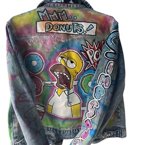 K&ouml;nnte beinhalten: Jeansjacke mit handgemaltem Design mit Homer Simpson und Donuts. Auf der R&uuml;ckseite der Jacke steht "Mmm.. Donuts." und auf dem &Auml;rmel "So Good". Die Jacke h&auml;ngt an einem wei&szlig;en Kleiderb&uuml;gel.