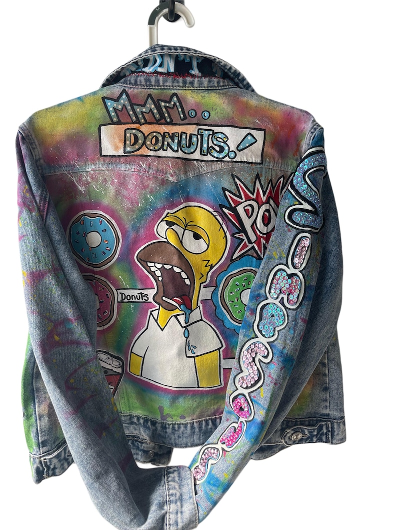 K&ouml;nnte beinhalten: Jeansjacke mit handgemaltem Design mit Homer Simpson und Donuts. Auf der R&uuml;ckseite der Jacke steht "Mmm.. Donuts." und auf dem &Auml;rmel "So Good". Die Jacke h&auml;ngt an einem wei&szlig;en Kleiderb&uuml;gel.