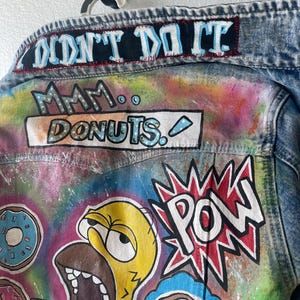 K&ouml;nnte beinhalten: Jeansjacke mit bunten Cartoon-Grafiken. Auf der R&uuml;ckseite der Jacke steht der Schriftzug "I DIDN'T DO IT" oben, mit "Mmm... Donuts" und einer Zeichentrickfigur mit gro&szlig;em offenem Mund. Das Wort "POW" wird ebenfalls angezeigt.
