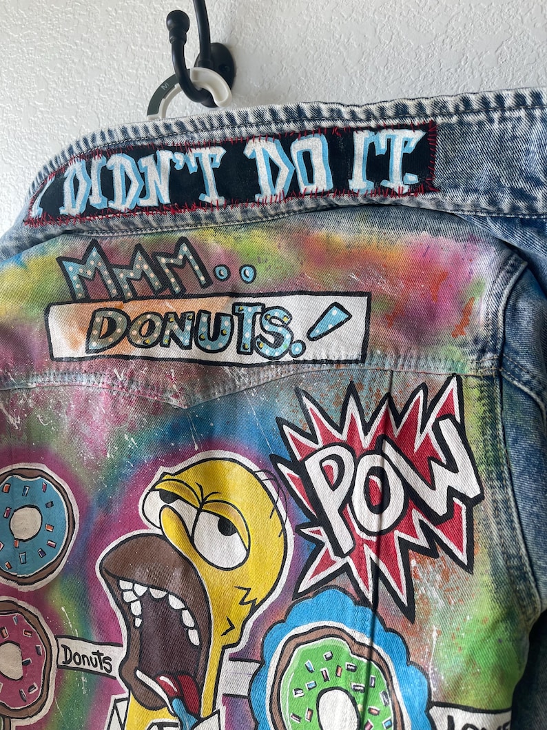 K&ouml;nnte beinhalten: Jeansjacke mit bunten Cartoon-Grafiken. Auf der R&uuml;ckseite der Jacke steht der Schriftzug "I DIDN'T DO IT" oben, mit "Mmm... Donuts" und einer Zeichentrickfigur mit gro&szlig;em offenem Mund. Das Wort "POW" wird ebenfalls angezeigt.