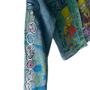 K&ouml;nnte beinhalten: Jeansjacke mit dem Wort "Someday" in Pailletten auf dem &Auml;rmel. Die Jacke ist mit bunten Farbspritzern und Strasssteinen verziert. Eine weitere Jacke mit einem Bart Simpson-Motiv ist sichtbar.