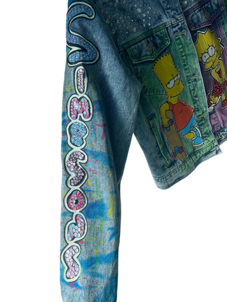 K&ouml;nnte beinhalten: Jeansjacke mit dem Wort "Someday" in Pailletten auf dem &Auml;rmel. Die Jacke ist mit bunten Farbspritzern und Strasssteinen verziert. Eine weitere Jacke mit einem Bart Simpson-Motiv ist sichtbar.