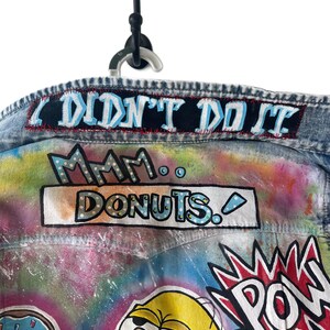 K&ouml;nnte beinhalten: Jeansjacke mit farbenfroher Graffiti-Kunst. Enth&auml;lt die Texte "I DIDN'T DO IT" und "Mmm... DONUTS." Enth&auml;lt Illustrationen von Zeichentrickfiguren und eine "POW"-Grafik. Die Jacke h&auml;ngt an einem schwarzen Haken.