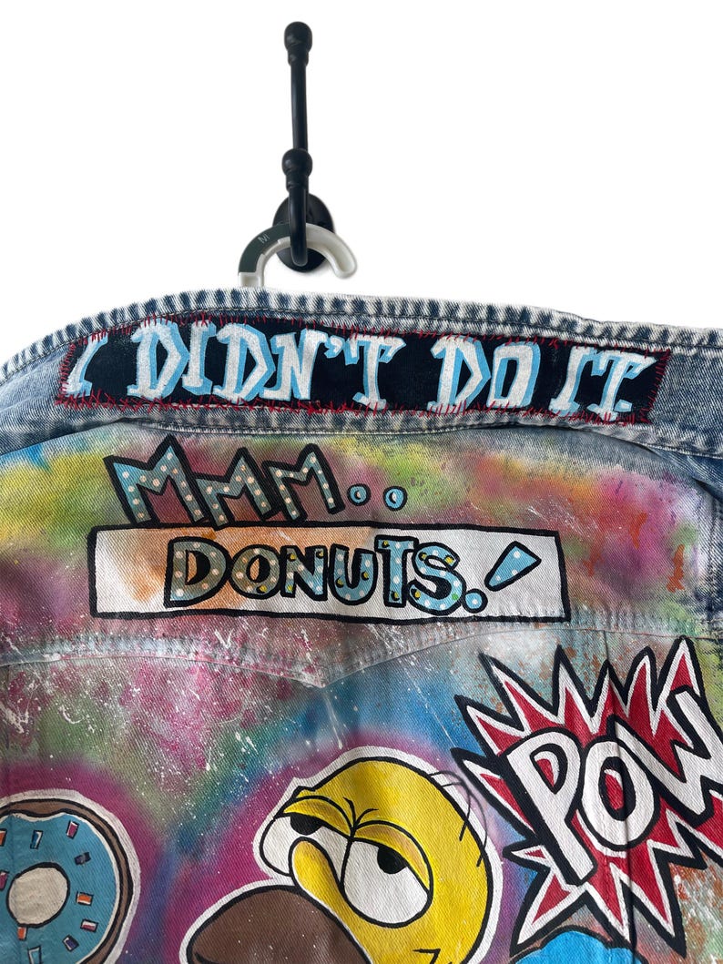 K&ouml;nnte beinhalten: Jeansjacke mit farbenfroher Graffiti-Kunst. Enth&auml;lt die Texte "I DIDN'T DO IT" und "Mmm... DONUTS." Enth&auml;lt Illustrationen von Zeichentrickfiguren und eine "POW"-Grafik. Die Jacke h&auml;ngt an einem schwarzen Haken.