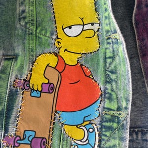 K&ouml;nnte beinhalten: Jeansjacke mit einem gro&szlig;en, aufgen&auml;hten Aufn&auml;her mit Bart Simpson aus der Zeichentrickserie Die Simpsons. Bart ist in seiner typischen Kleidung dargestellt und h&auml;lt ein Skateboard. Die Jacke hat einen gr&uuml;n-blauen Batik-Effekt.