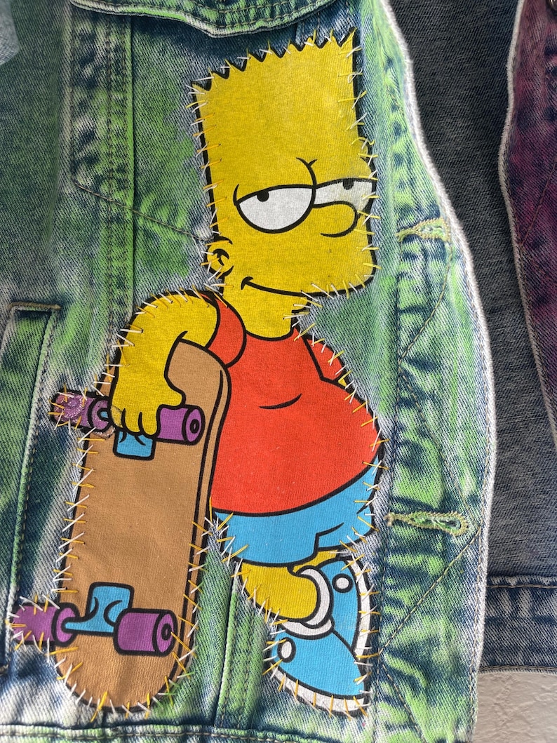 K&ouml;nnte beinhalten: Jeansjacke mit einem gro&szlig;en, aufgen&auml;hten Aufn&auml;her mit Bart Simpson aus der Zeichentrickserie Die Simpsons. Bart ist in seiner typischen Kleidung dargestellt und h&auml;lt ein Skateboard. Die Jacke hat einen gr&uuml;n-blauen Batik-Effekt.