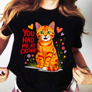 Pode incluir: T-shirt preto com um desenho de gato tigrado laranja de desenho animado, com olhos verdes e listras vermelhas. O texto diz "You had me at Catnip". O design inclui cora&ccedil;&otilde;es e flores coloridas, criando um design divertido e l&uacute;dico.