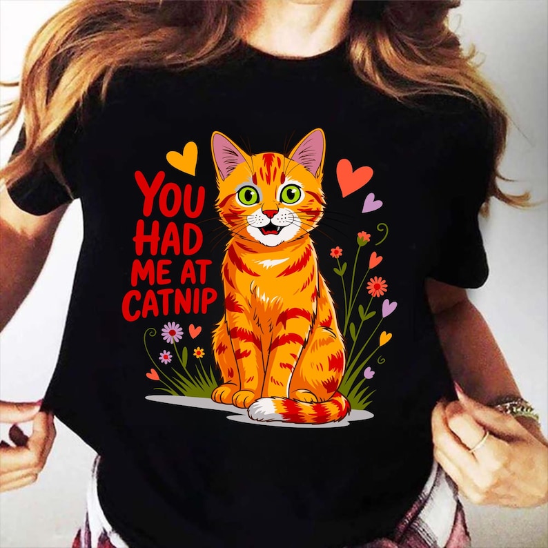Pode incluir: T-shirt preto com um desenho de gato tigrado laranja de desenho animado, com olhos verdes e listras vermelhas. O texto diz "You had me at Catnip". O design inclui cora&ccedil;&otilde;es e flores coloridas, criando um design divertido e l&uacute;dico.