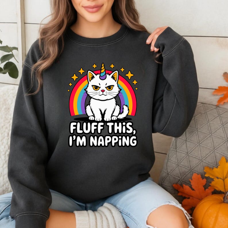 Pode incluir: Camisola cinza escuro com um gato branco com um chifre de unic&oacute;rnio, contra um arco-&iacute;ris e estrelas. O texto "FLUFF THIS, I'M NAPPING" est&aacute; impresso abaixo. Uma parte de cima casual e confort&aacute;vel.
