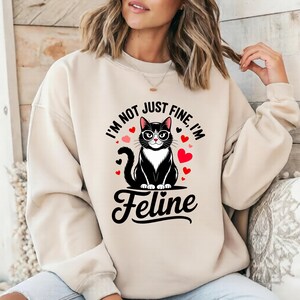 Pode incluir: Camisola bege com um gato preto de &oacute;culos, rodeado por cora&ccedil;&otilde;es vermelhos. O texto diz "I'm not just fine, I'm Feline". O gato tem o peito branco e est&aacute; sentado em posi&ccedil;&atilde;o vertical. Um design fofo para os amantes de gatos.