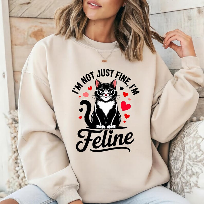 Pode incluir: Camisola bege com um gato preto de &oacute;culos, rodeado por cora&ccedil;&otilde;es vermelhos. O texto diz "I'm not just fine, I'm Feline". O gato tem o peito branco e est&aacute; sentado em posi&ccedil;&atilde;o vertical. Um design fofo para os amantes de gatos.