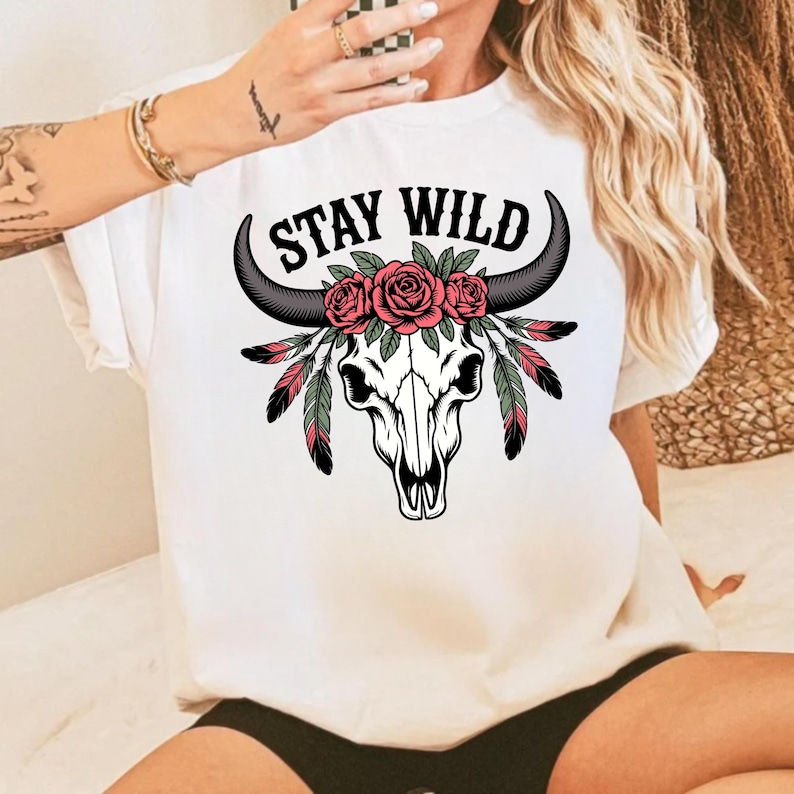 May include: White t-shirt with a bull skull graphic and the words "Stay Wild". The skull is decorated with pink roses, black horns, and feathers in shades of green and red. The design is centered on the front.