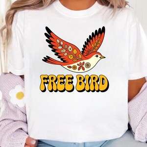 May include: White t-shirt with a retro-style bird graphic in orange, red, and yellow. The bird is flying above the text "FREE BIRD" in a bold, yellow font with a black outline.