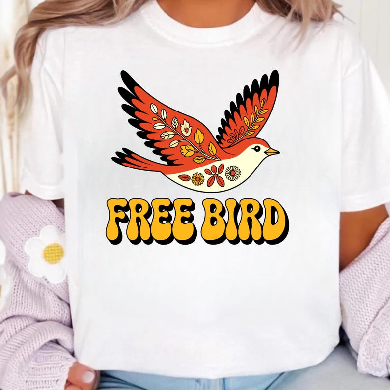 May include: White t-shirt with a retro-style bird graphic in orange, red, and yellow. The bird is flying above the text "FREE BIRD" in a bold, yellow font with a black outline.