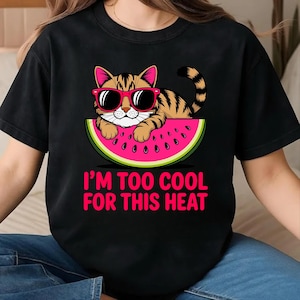 Pode incluir: Camiseta preta com um gato de desenho animado usando &oacute;culos de sol, descansando em uma fatia de melancia. O gato tem listras laranja e marrom. O texto "I'M TOO COOL FOR THIS HEAT" &eacute; impresso em rosa.
