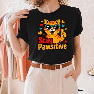 Pode incluir: Camiseta preta com um gato tabby laranja de desenho animado usando &oacute;culos de sol e com o polegar para cima. O texto "Stay Pawsitive" est&aacute; em amarelo e vermelho. O design inclui cora&ccedil;&otilde;es e pegadas. Design divertido.